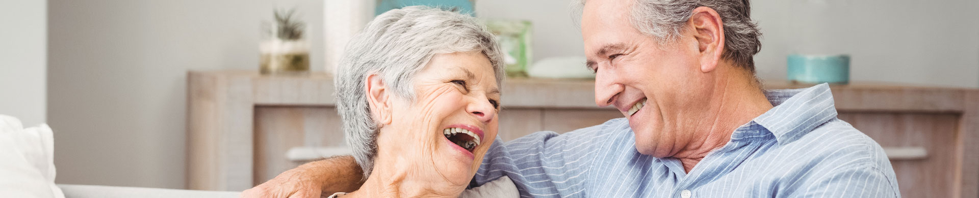 A senior man wraps his arms around a senior woman and they both laugh the smile at each other
