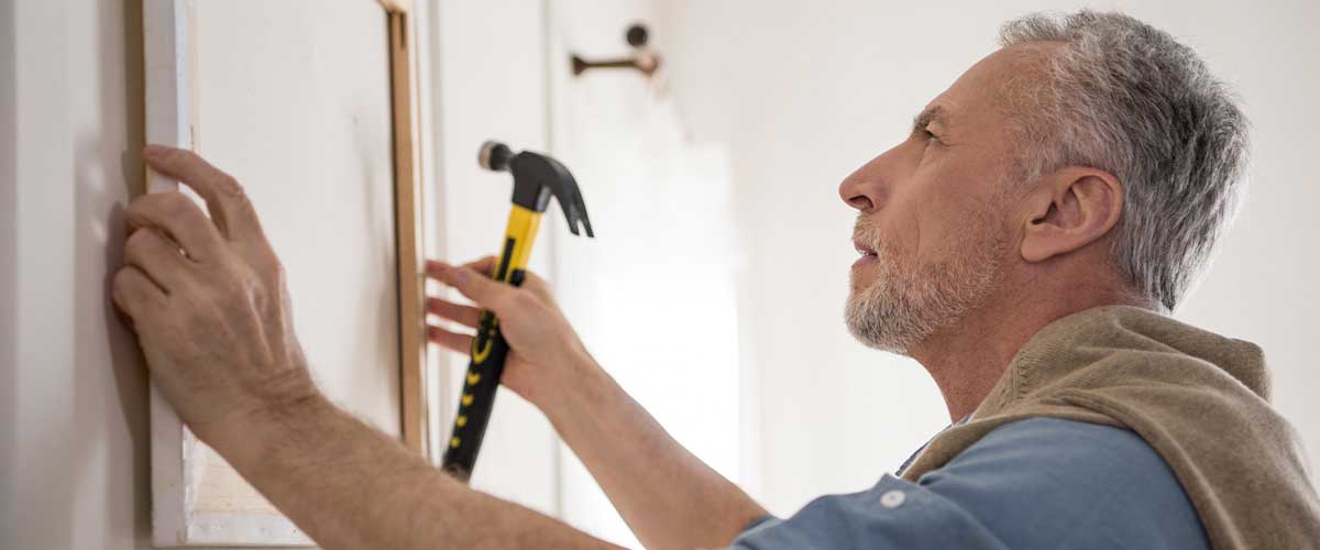 A senior man holds a hammer as he positions a framed photo on the wall