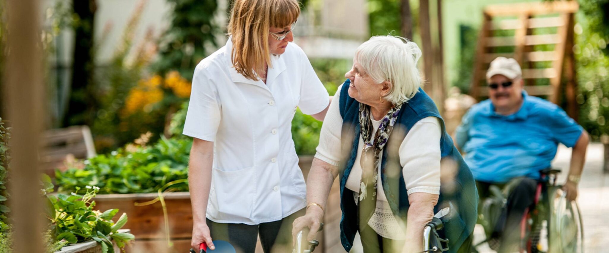 A senior woman moves with the help of walker and a health care professional and behind her on her outdoor walk is a man in a wheelchair