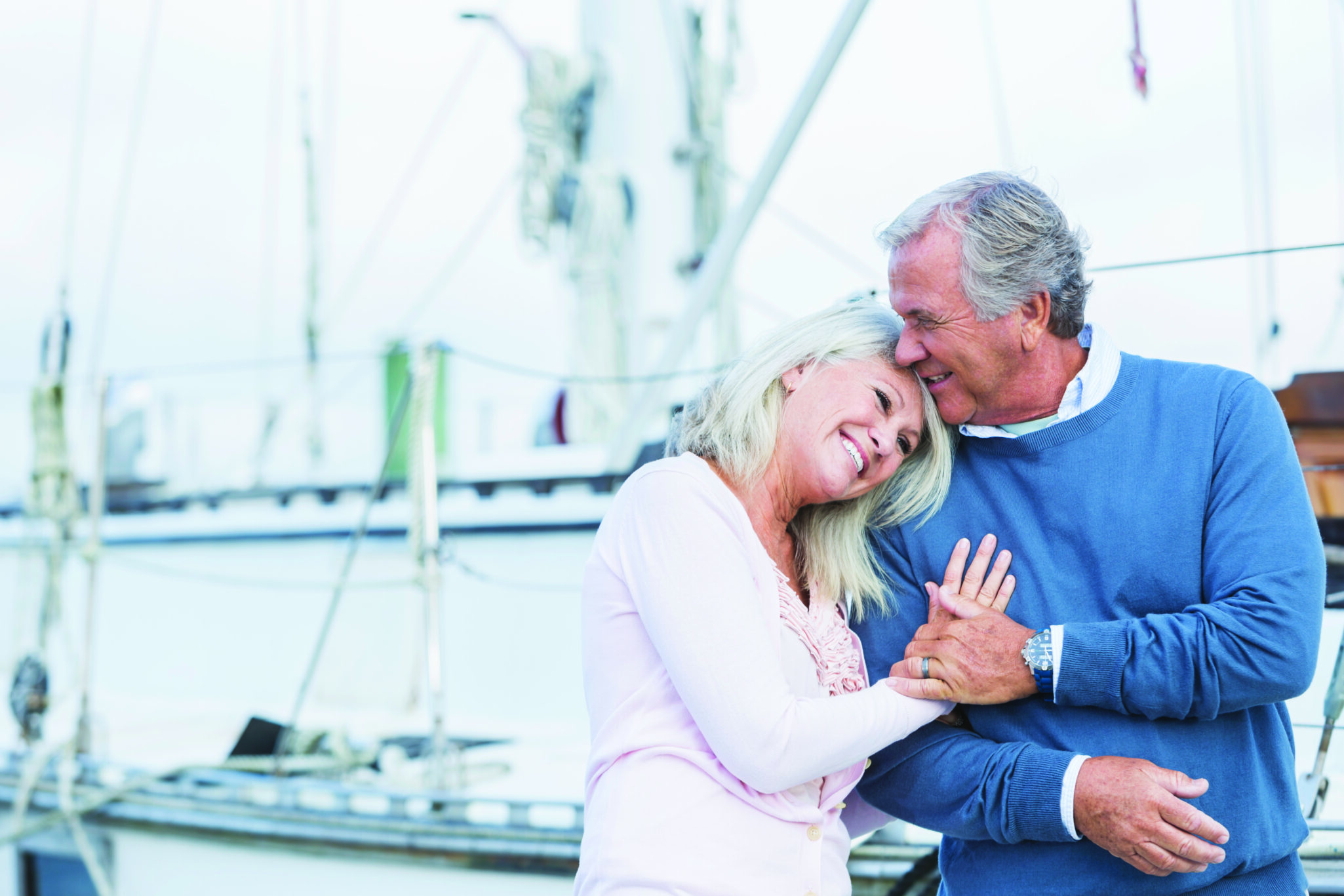 An affectionate, mature couple at a marina standing together on a dock, luxury boats and yachts in the background. The senior man is holding his wife's hand, and she is resting her head on his shoulder, smiling.
