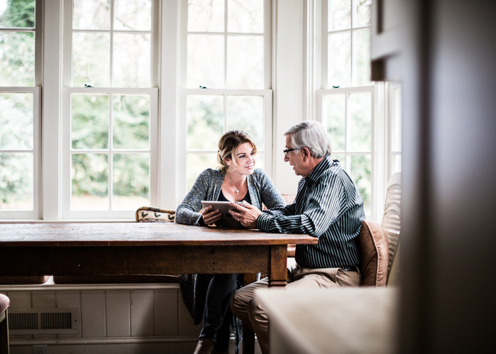 A woman sits at the table with her older father and and they look at a tablet together